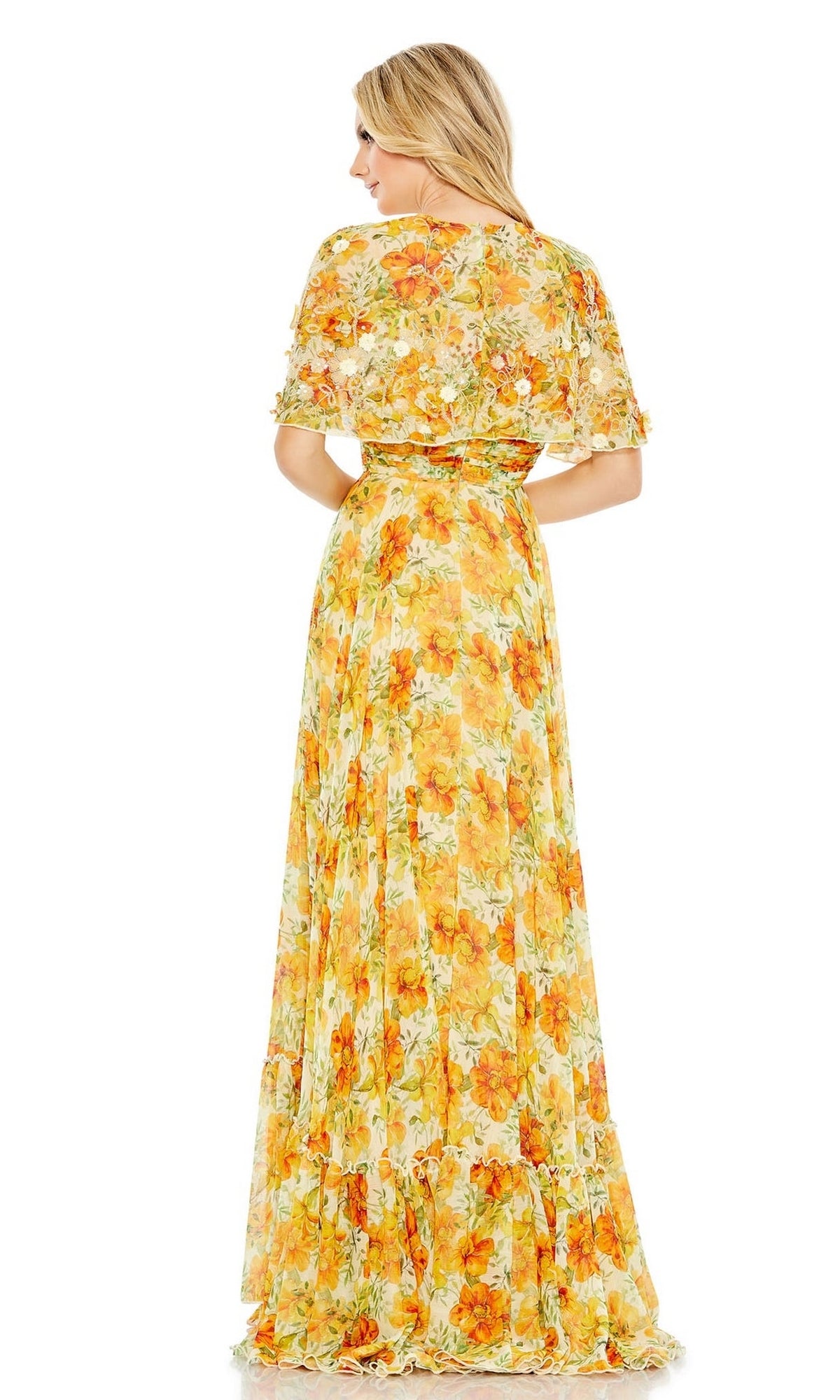 A woman stands facing away, wearing the Long Formal Dress 9201 by Mac Duggal—a floor-length, yellow floral A-line cape dress with short sleeves and a flowing skirt.